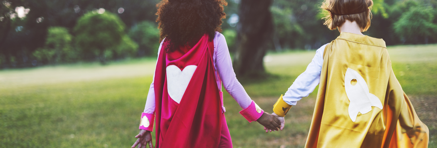 Two kids wearing super hero costumes holding hands in a field